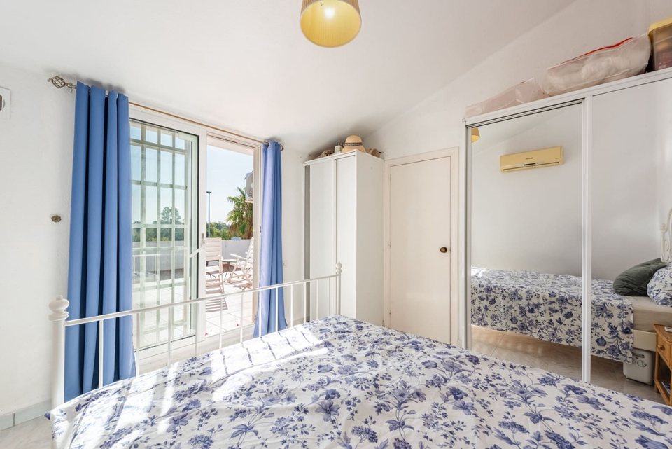 Dormitorio con cama de matrimonio blanca y colcha floral, cortinas claras y mesilla