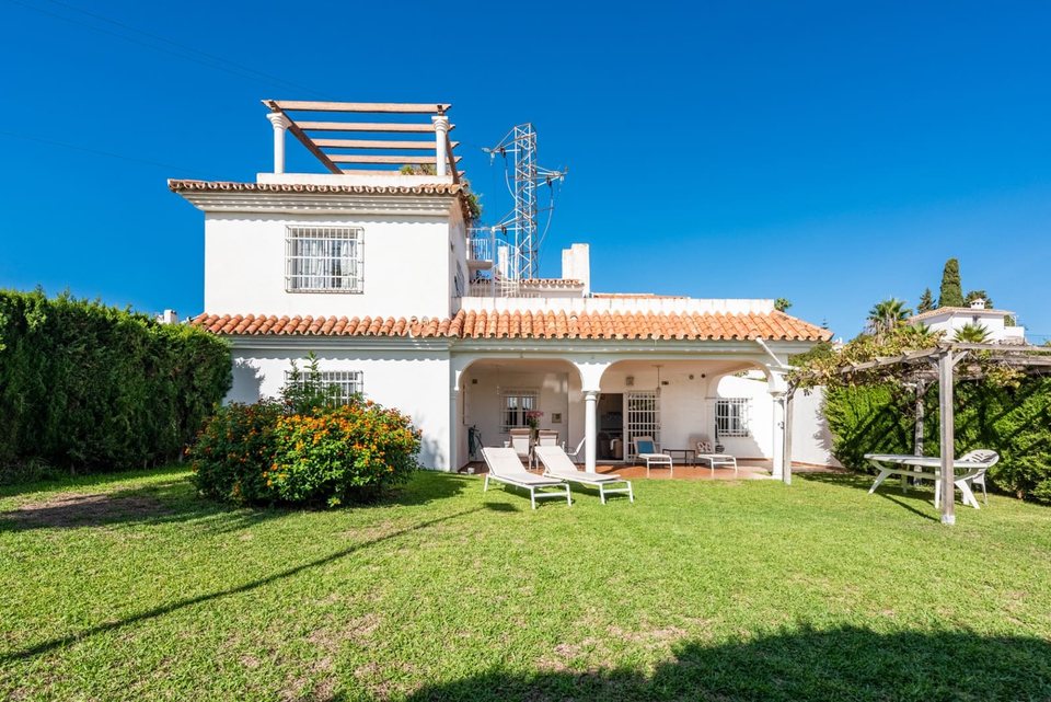 Fachada frontal del chalet con jardín llano, setos verdes y tumbonas blancas