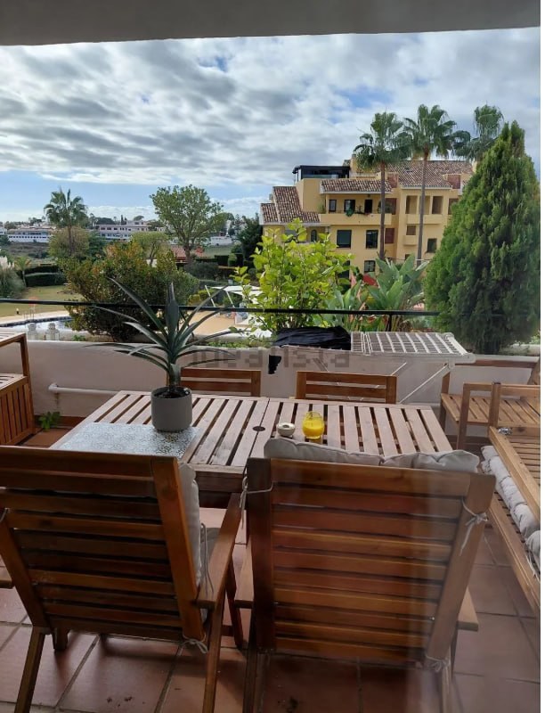 Terraza principal del piso con mesa de madera, sillas y vistas abiertas al mar y a los tejados del conjunto