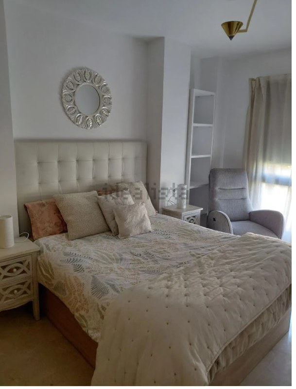 Dormitorio principal con cabecero tapizado en gris claro, mesitas de noche blancas, espejo redondo decorativo y butaca gris
